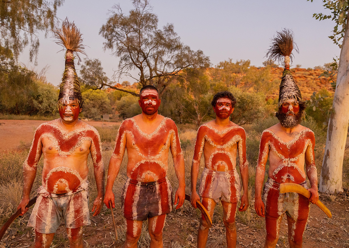 Four Aboriginal men with red and white body paint designs, ceremonial headwear and boomerangs among trees 