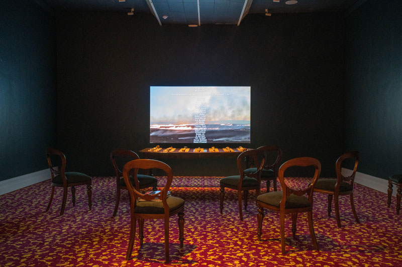 Julie Gough, The Gathering, 2015, 2019, HDMI video, sound, edited by Jemma Rea, colonial table, enamel on Tasmanian oak, 28 found stones, carpet; Julie Gough with Margaret Woodward, Entitled (red), 2019, designer Jet® bespoke carpet, custom made by Godfrey-Hirst Australia. Installation view, Julie Gough: Tense Past, Tasmanian Museum and Art Gallery. Photo: Alastair Bett